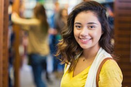 Pretty trendy Hispanic female high school or college student is standing in modern library. She is wearing a yellow t-shirt with a beige vest. She is looking at the camera and smiling. People are in the background searching for books.